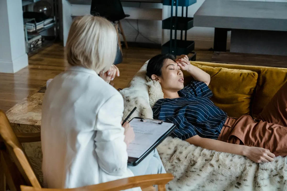 A counselor with a clipboard sits beside a person resting on a couch with a hand raised to their forehead.