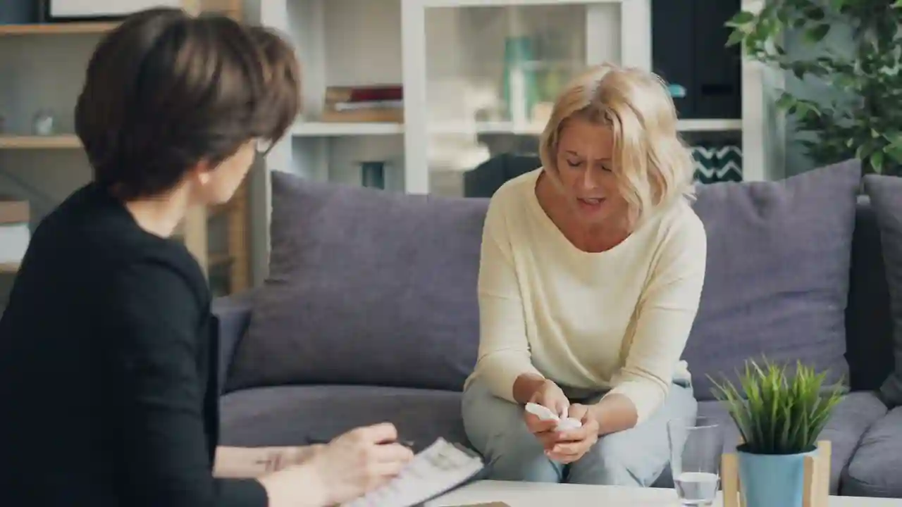 A woman holding a tissue speaks tearfully with a counselor who takes notes in a bright sitting room.